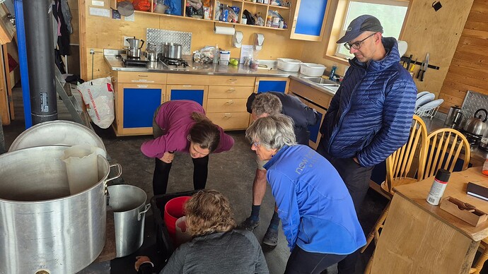 People standing around watching a stove. Wise custodians find inside work when the weather gets poor. Here we learn about fixing the pellet stove.